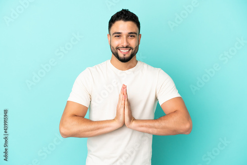 Young arab man isolated on blue background keeps palm together. Person asks for something