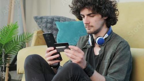 Side view of a curly haired young man shopping online with credit card using smart phone at home. Enter the card details to pay for the online purchase. photo