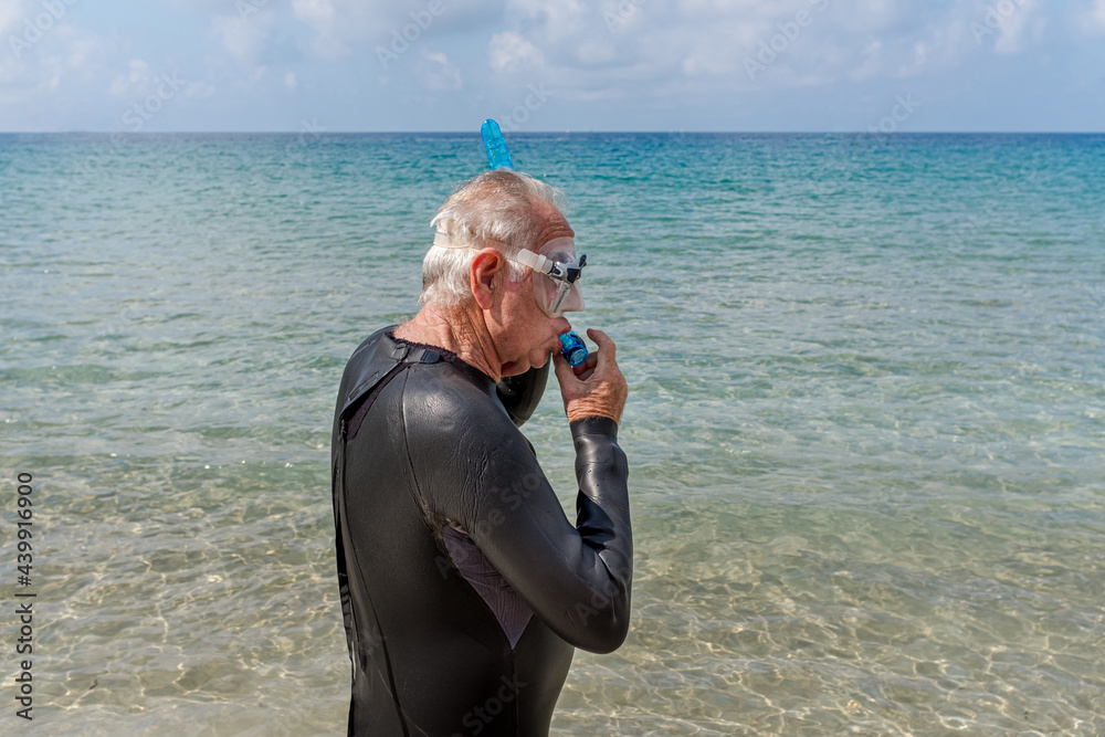 Older retired man on the beach wearing a wetsuit and putting on his snorkel goggles to go swimming.