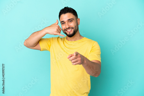 Young arab man isolated on blue background making phone gesture and pointing front