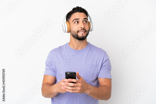 Young arab man isolated on white background listening music with a mobile and thinking