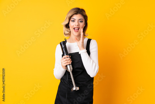 Girl using hand blender isolated on yellow background with surprise and shocked facial expression