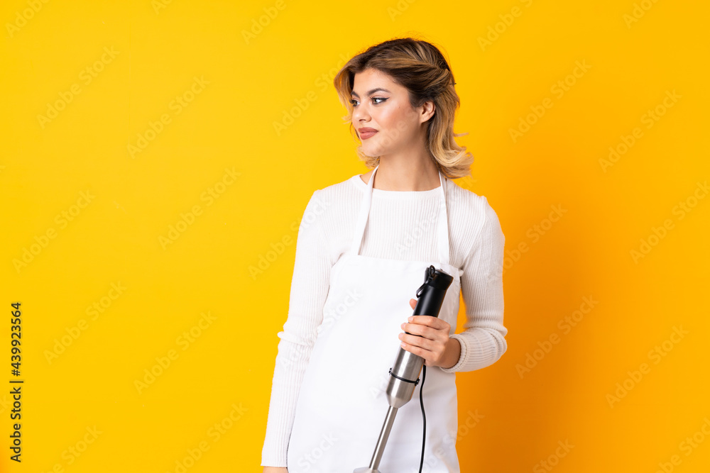 Girl using hand blender isolated on yellow background looking to the side and smiling