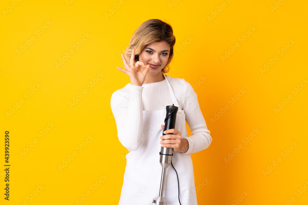 Girl using hand blender isolated on yellow background showing ok sign and thumb up gesture Girl using hand blender isolated on yellow background showing ok sign and thumb up gesture