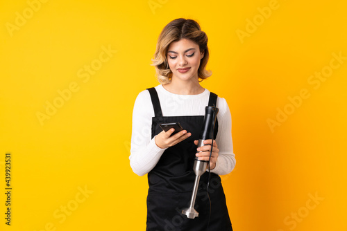 Girl using hand blender isolated on yellow background sending a message with the mobile © luismolinero