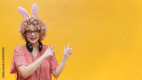 Excited young girl wearing bunny ears and glasses pointing at something. Girl in joyful mood. Headphones around her neck. Dressed in pink shirt. Isolated over yellow background studio. 