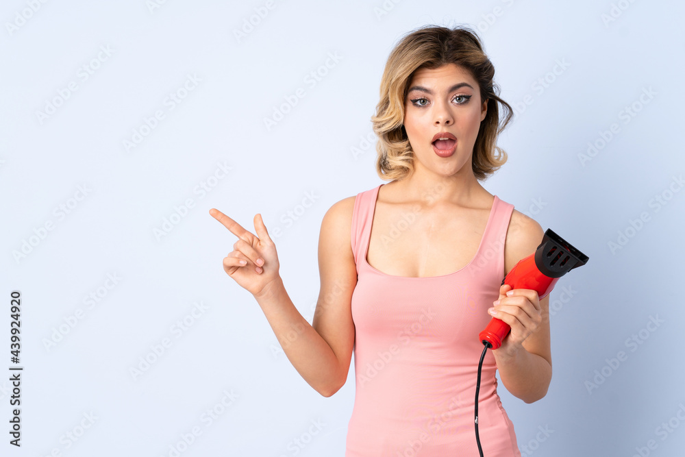 Russian girl holding a hairdryer isolated on blue background surprised and pointing side Russian girl holding a hairdryer isolated on blue background surprised and pointing side