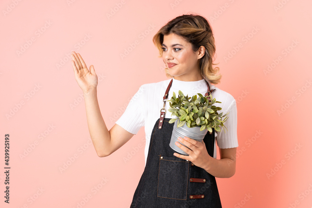 Russian gardener girl holding a plant isolated on pink background extending hands to the side for inviting to come Russian gardener girl holding a plant isolated on pink background extending hands to the side for inviting to come