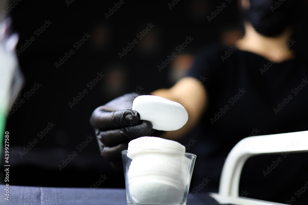 the tattoo artist holds a cotton pad in his hand. girl master of permanent makeup lies a cotton sponge in a glass