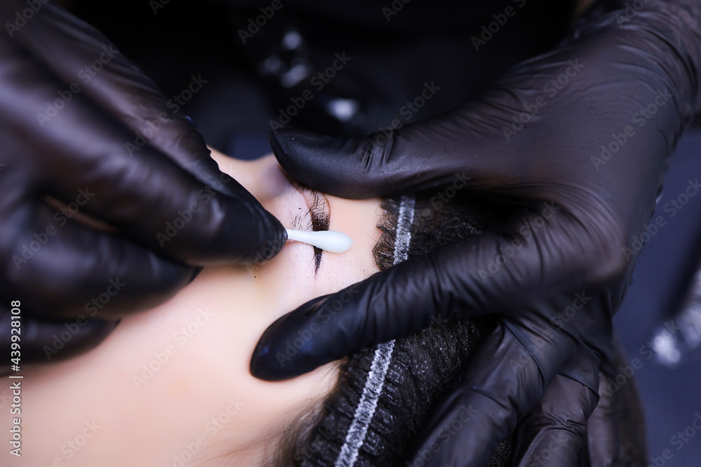 the tattoo artist's hands close up with one hand holding the client's eyebrows and stretching it with the other hand holding a cotton swab and wiping away the remaining pigment