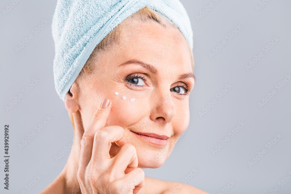 Senior smiling 50s middle aged mature older woman applying facial cream on face looking at camera isolated over white background. Anti age healthy dry skin care beauty therapy concept Senior smiling 50s middle aged mature older woman applying facial cream on face looking at camera isolated over white background. Anti age healthy dry skin care beauty therapy concept