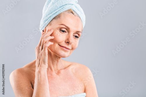 Senior smiling 50s middle aged mature older woman applying facial cream on face looking at camera isolated over white background. Anti age healthy dry skin care beauty therapy concept