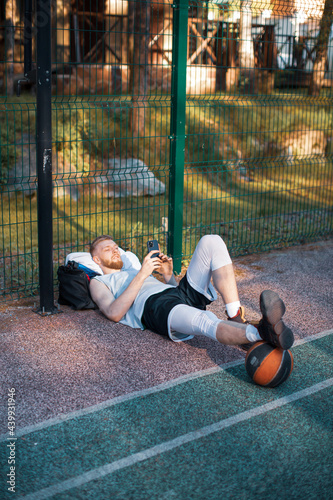 Basketball player sportive guy lying relaxing with phone after workout outdoors on basketball court