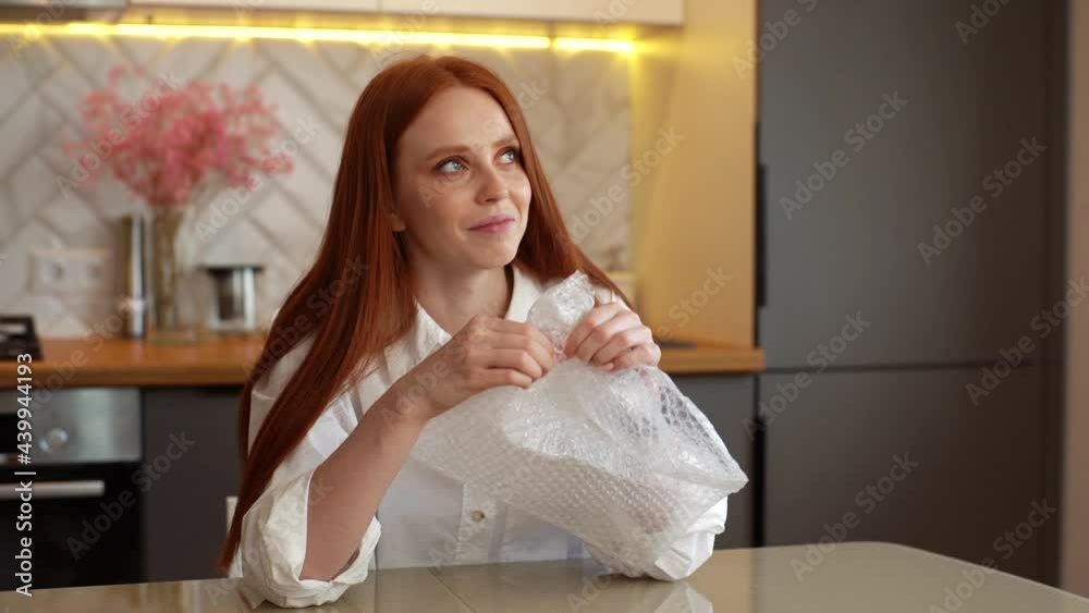 Middle shot portrait of happy young woman playing pops bubble wrap to calm herself sitting at table in kitchen room. Smiling redhead female popping plastic bubblewrap. Shooting in slow motion. Middle shot portrait of happy young woman playing pops bubble wrap to calm herself sitting at table in kitchen room. Smiling redhead female popping plastic bubblewrap. Shooting in slow motion.