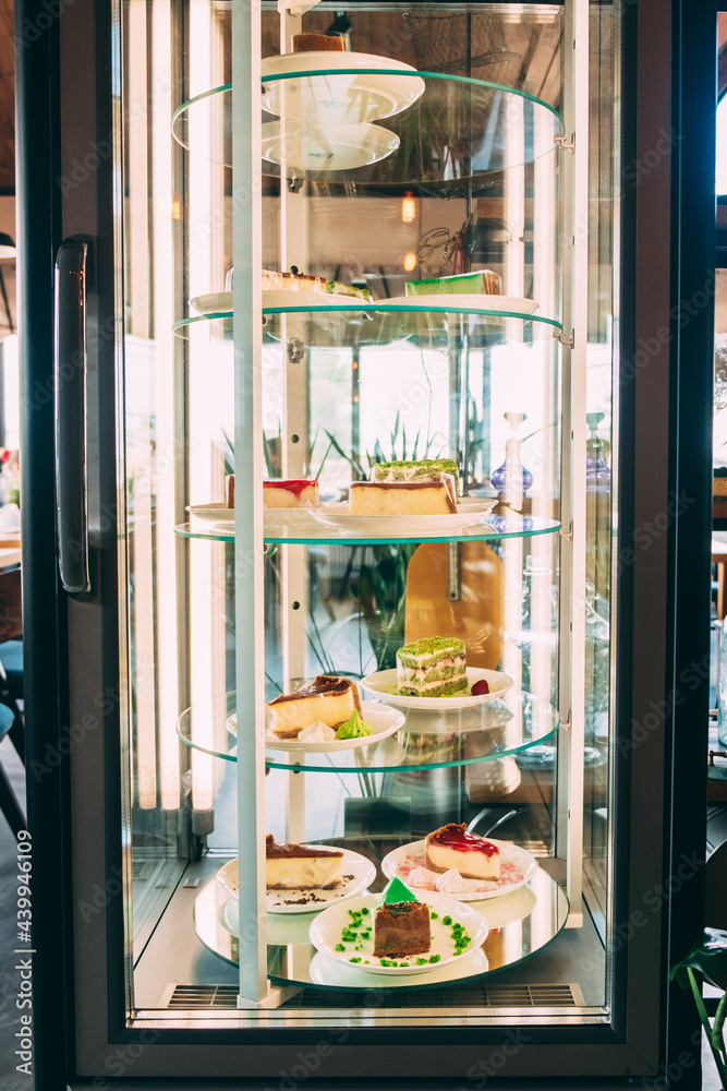 Glass showcase refrigerator with desserts inside illuminated by lamps.