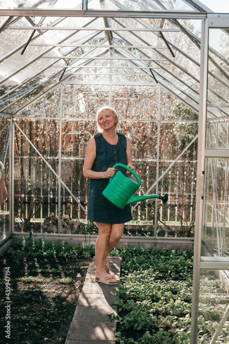 female gardener is watering plants in greenhouse. She holds a plastic green watering can.