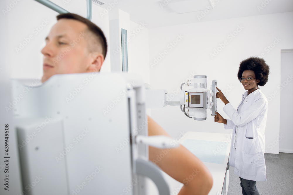 Young man standing in modern hospital during x ray chest scan, while female Afro American medical technician works with X-Ray machine. Scanning for fractures, chest, cancer or tumor Young man standing in modern hospital during x ray chest scan, while female Afro American medical technician works with X-Ray machine. Scanning for fractures, chest, cancer or tumor