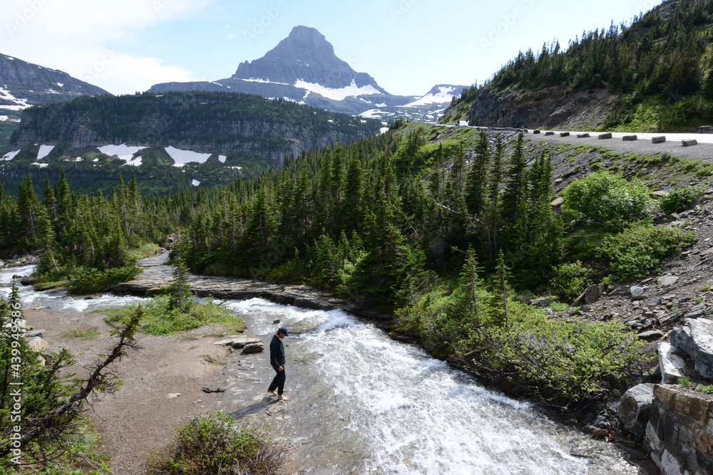 A man taking a closer look at a river in Glacier National Park, with snow capped mountains in the background