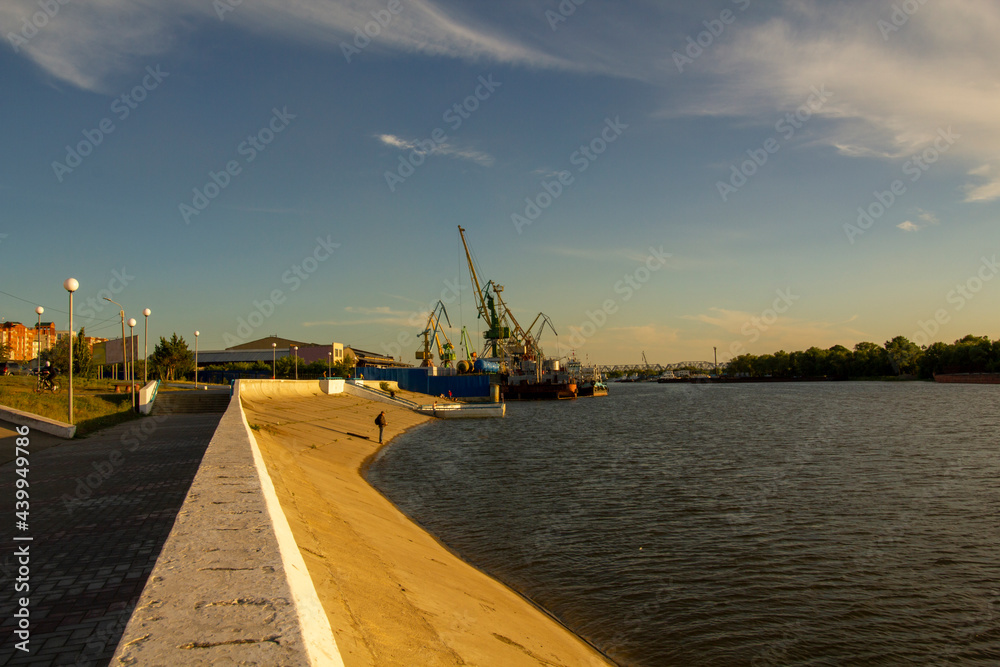 View from the river embankment on the river port. Port cranes and the embankment of the river in the rays of the sunset.