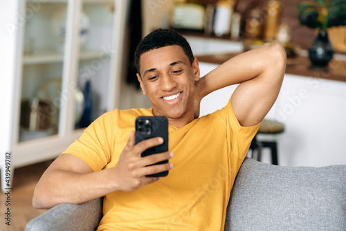 Satisfied joyful hispanic guy dressed in a yellow t-shirt, sitting in living room on a sofa, holding and using his smartphone, texting online, browsing social networks, looking at phone, smiling photo