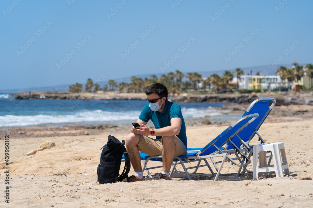 Tourism during pandemic. Safe traveling. Coronavirus travel ban. Quarantine due coronavirus epidemic. Sick tired young man in face mask sit on deck chair on beach by mediterranean sea in city Paphos