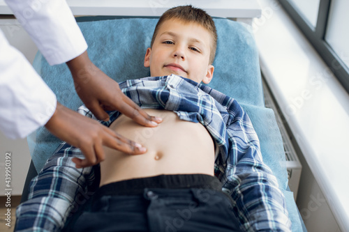 Cute smiling 10-aged kid boy, lying on the couch at modern clinic, visiting his doctor. Female afro pediatrician examining abdomen of child patient using manual palpation photo