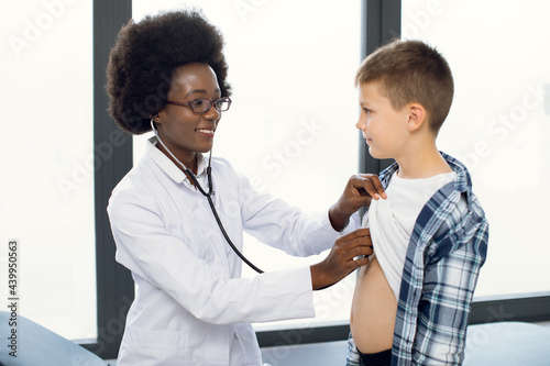 Cute likable Caucasian teen boy and joyful African woman doctor, during doctors checkup. Pediatrist examinates young patient's heartbeat and lungs with stethoscope