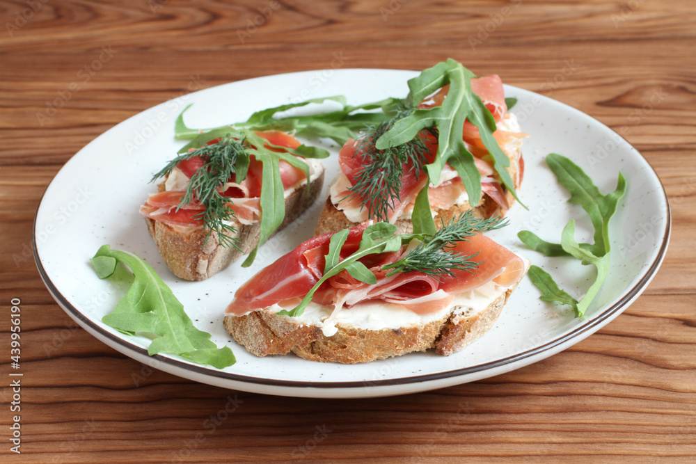 Buckwheat toasts with goat cheese, ham and arugula lying on a white plate on a wooden table.  Closeup