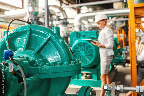 Successful smiling hardworking female manager in suit standing in heating plant with tablet in hands and checking on turbine.