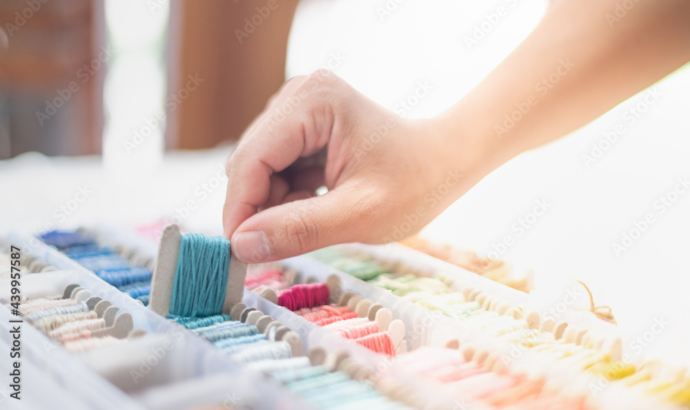 Close up hands of tailor woman holding ocean blue threads and different colors in the box put on the table and prepare to use with white cotton and frame wood. Close up hands of tailor woman holding ocean blue threads and different colors in the box put on the table and prepare to use with white cotton and frame wood.