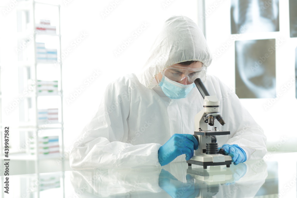 Doctor in a protective medical suit wearing a mask wearing blue gloves in a laboratory looks through a microscope