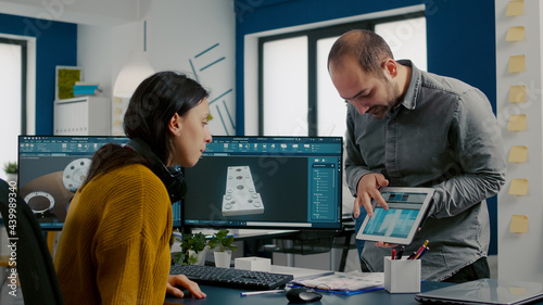 Industrial designer discussing with colleague holding tablet with in CAD program, designing 3D prototype of building. Woman engineer using pc with two monitors sharing ideas for construction mechanism photo