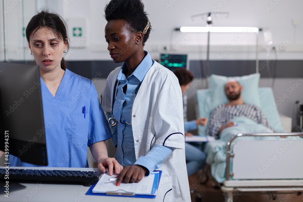 Fototapeta premium Woman nurse explaining medical recovery on computer to afro american practitioner in hospital ward. Hospitalized man patient discussing with doctor illness treatment during expertise consultation