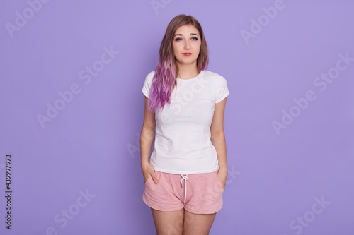 Young beautiful sad and pensive female looking at camera, standing with hands in pockets, wearing white casual t-shirt and pink shirts, isolated over lilac background.