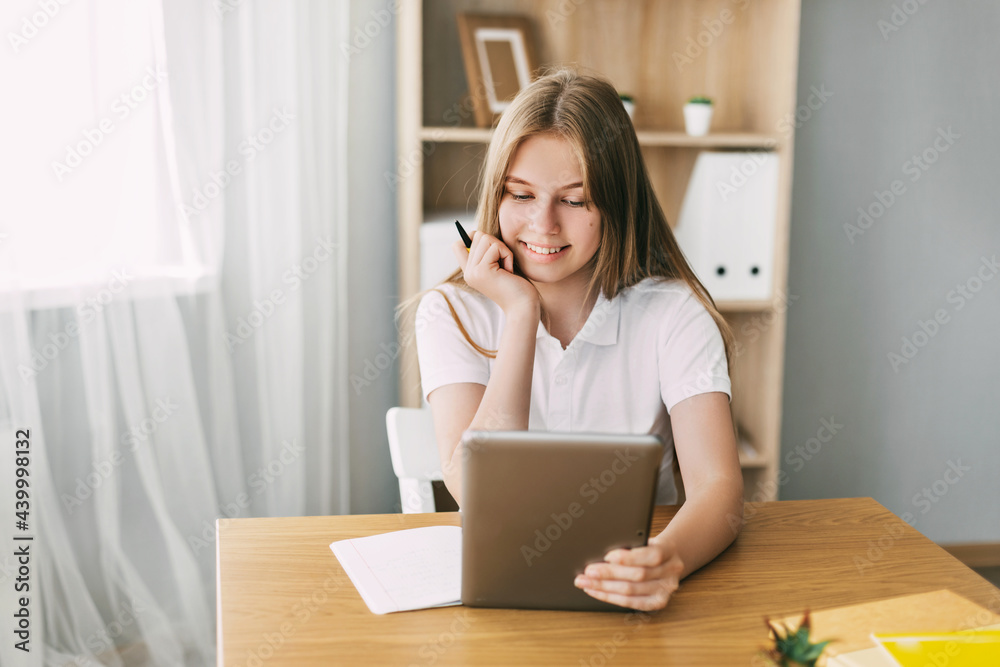 A thoughtful girl sits at a desk and prepares for exams using a tablet A thoughtful girl sits at a desk and prepares for exams using a tablet