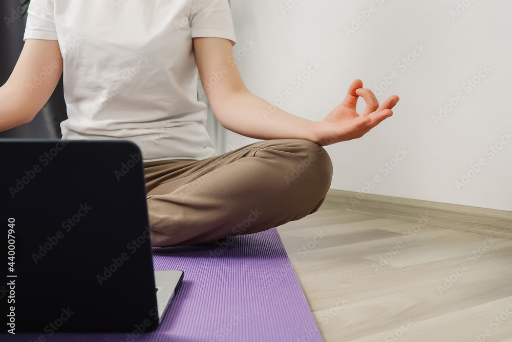 Unrecognizable female meditating in lotus position sitting, watch online class tutorial on laptop at home. Woman doing morning practice in living room. Hand of women in lotus pose while sit at floor