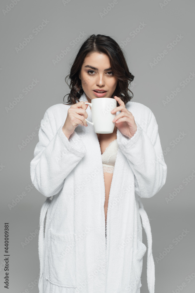 Pretty woman in bra and bathrobe holding cup isolated on grey Pretty woman in bra and bathrobe holding cup isolated on grey