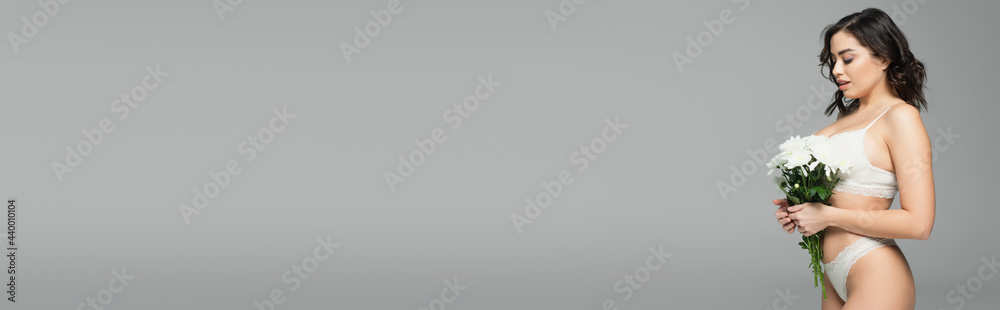 Side view of pretty woman in lingerie looking at flowers isolated on grey, banner