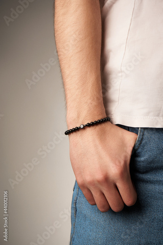 Cropped close-up shot of male wrist with beaded bracelet made of black ornamental stone and decorated with metal beads and charm with letter B. Man in beige t-shirt put his hand in the pocket.  photo