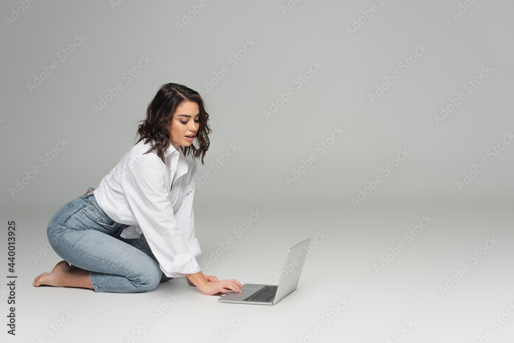 Pretty woman in white shirt and jeans using laptop on grey background Pretty woman in white shirt and jeans using laptop on grey background