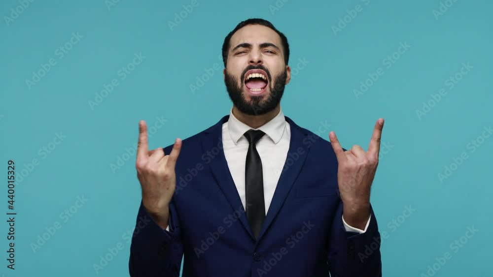 Enthusiastic crazy brunette guy in formal suit showing rock and roll sign gesture with fingers up and making grimaces, yelling happily. Indoor studio shot isolated on blue background. Enthusiastic crazy brunette guy in formal suit showing rock and roll sign gesture with fingers up and making grimaces, yelling happily. Indoor studio shot isolated on blue background.
