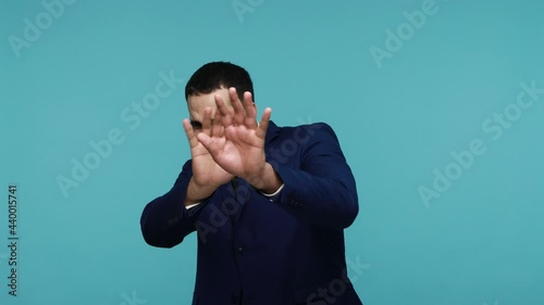 Worried scared brunette man in black suit raising his hands in defensive gesture and looking in horror at camera, walking away from frightening thing. Indoor studio shot isolated on blue background.