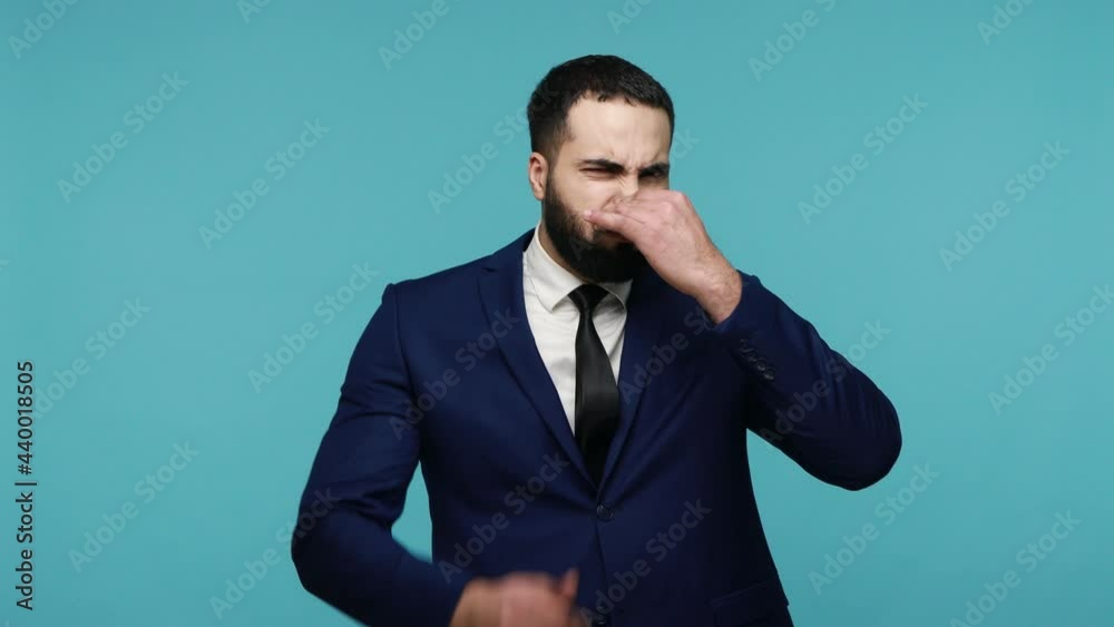 Confused disgusted handsome man with beard in suit pinching nose from bad awful smell, frowning in displeasure, showing stop with raised hand. Indoor studio shot isolated on blue background. Confused disgusted handsome man with beard in suit pinching nose from bad awful smell, frowning in displeasure, showing stop with raised hand. Indoor studio shot isolated on blue background.