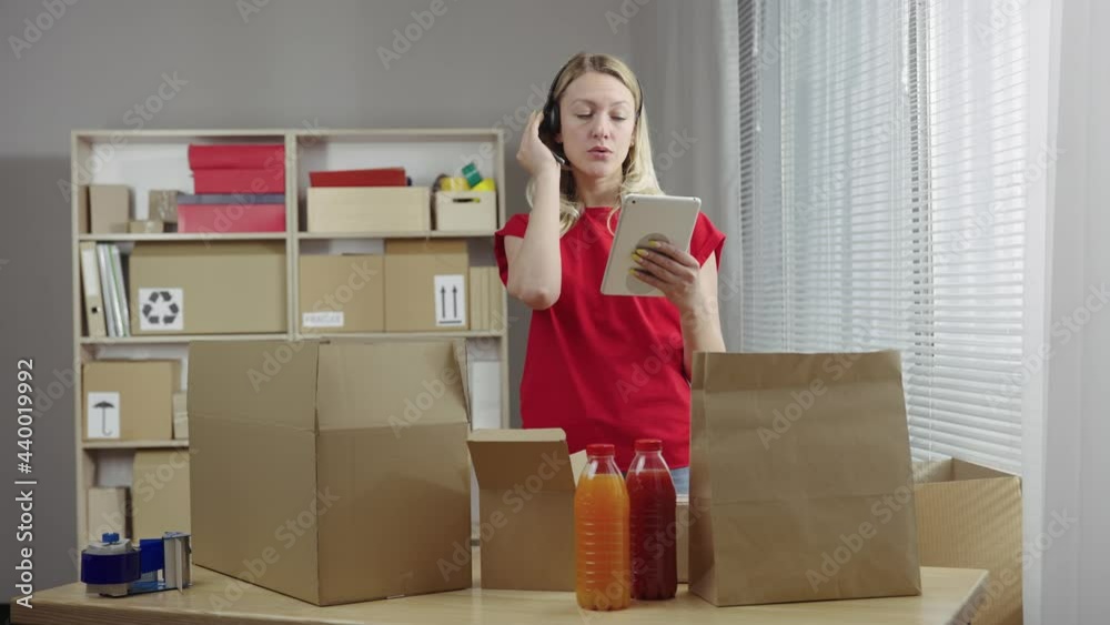 Warehouse worker is working in an office room against background of boxes. Woman prepares parcels of food for delivery to customers using a headset and a tablet. Close up. Slow motion ready 59.94fps. Warehouse worker is working in an office room against background of boxes. Woman prepares parcels of food for delivery to customers using a headset and a tablet. Close up. Slow motion ready 59.94fps.