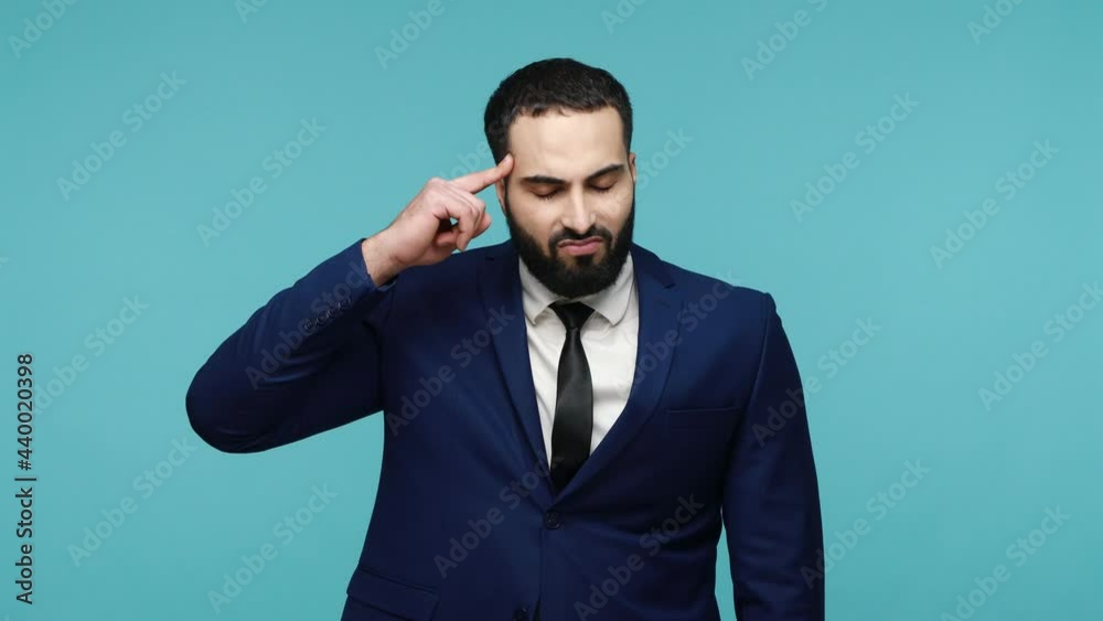 Displeased upset brunette bearded man in formal suit making stupid gesture with finger near head, looking down sadly, frustrated depressed expression. Indoor studio shot isolated on blue background. Displeased upset brunette bearded man in formal suit making stupid gesture with finger near head, looking down sadly, frustrated depressed expression. Indoor studio shot isolated on blue background.