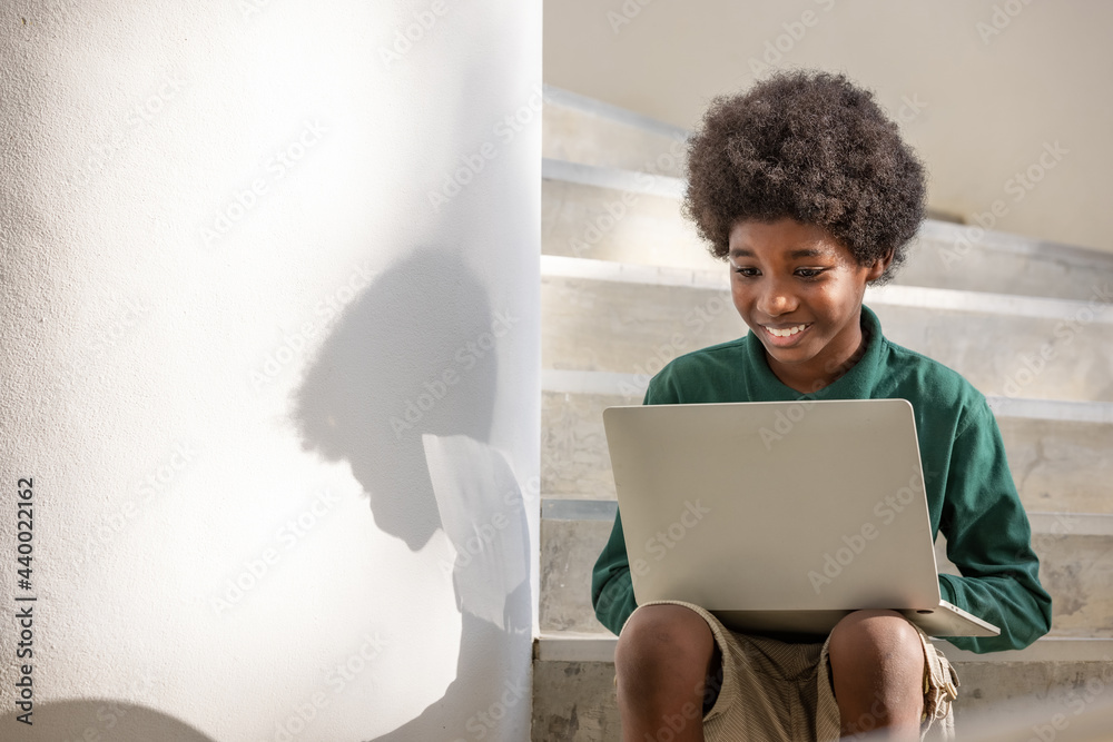 An African curly haired boy sits on a laptop in the corridor downstairs under a building. An African curly haired boy sits on a laptop in the corridor downstairs under a building.