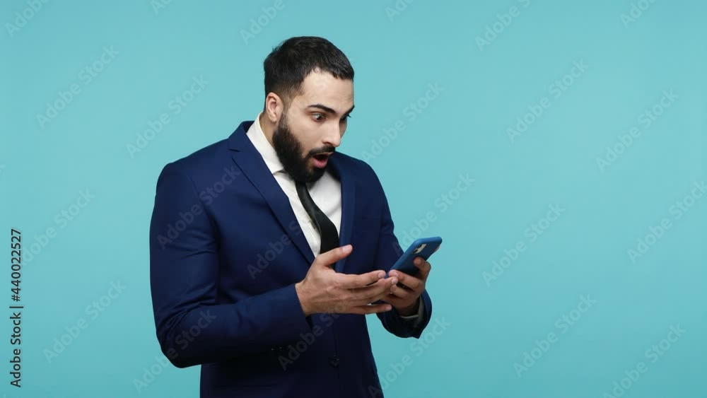 Shocked bearded businessman in formal suit looks surprisingly and with terrified expression at screen of smart phone, reads breaking news via internet.,Indoor studio shot isolated on blue background. Shocked bearded businessman in formal suit looks surprisingly and with terrified expression at screen of smart phone, reads breaking news via internet.,Indoor studio shot isolated on blue background.