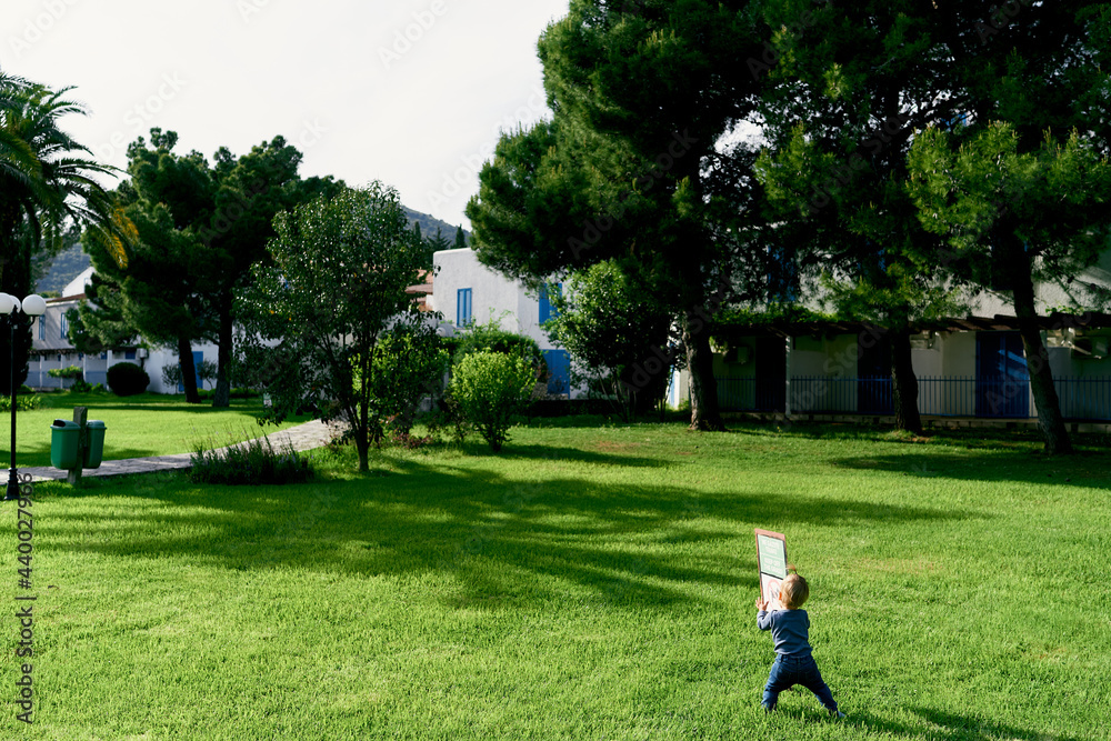 Kid stands on a green lawn near a signpost against the background of trees and houses