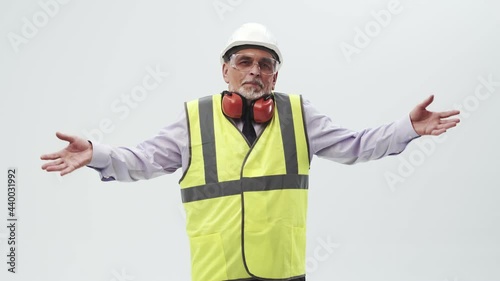 Adult man engineer in confusion spreads hands to the sides, standing in a vest and helmet with headphones on a white studio background, looking into the camera. Concept of ignorance and unknown.