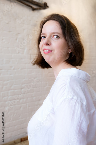 Vertical portrait of young woman with shoulder-length dark hair in white shirt on white brick wall background. Woman's portrait to the waist in a half turn. Woman smiling